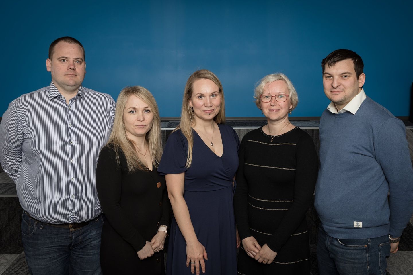 Ryhmäkuva viidestä ihmisestä, jotka seisovat yhdessä sisätiloissa sinistä seinää vasten.