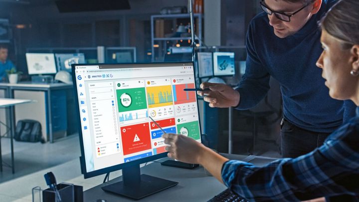 Two people analyze a computer screen displaying a dashboard with charts, graphs, and alerts in an office setting.